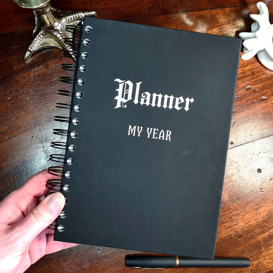 A Dateless Gothic Planner with a black hardback cover that says Planner and My Year on the front in white.