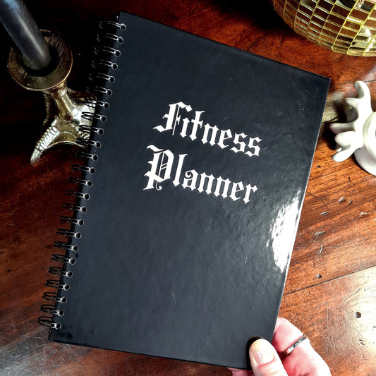 A Gothic Fitness Planner with a black hardback cover that says Fitness Planner in white font.